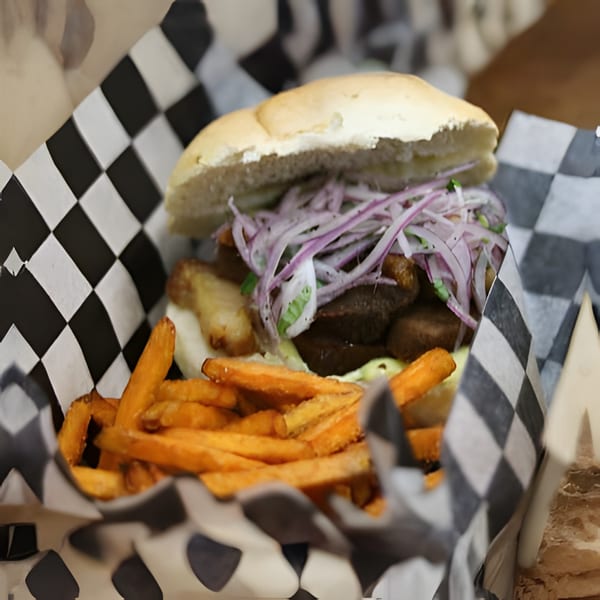 A sandwich with meat, onions, and sauce, served with sweet potato fries in a checkered basket.
